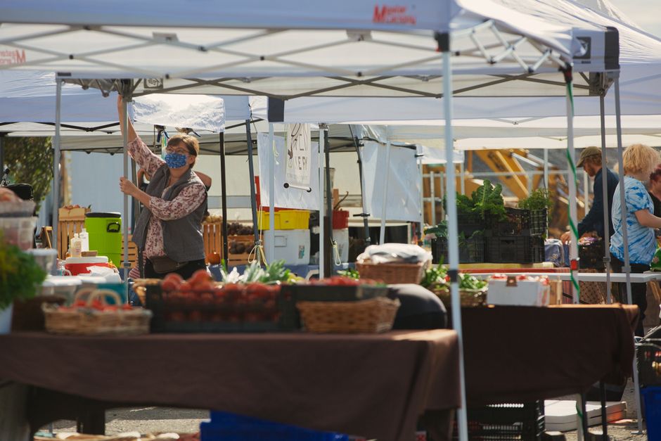 A woman wearing a floral dress and a face mask is standing behind a market stall, adjusting a canopy tent at an outdoor farmers' market. The stall features various fresh produce displayed in baskets,