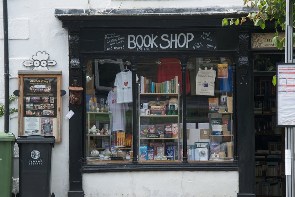 The image shows the front of a quaint bookshop with a black-painted facade, featuring large glass windows displaying a variety of items including a white t-shirt with a red heart, books, and small dec