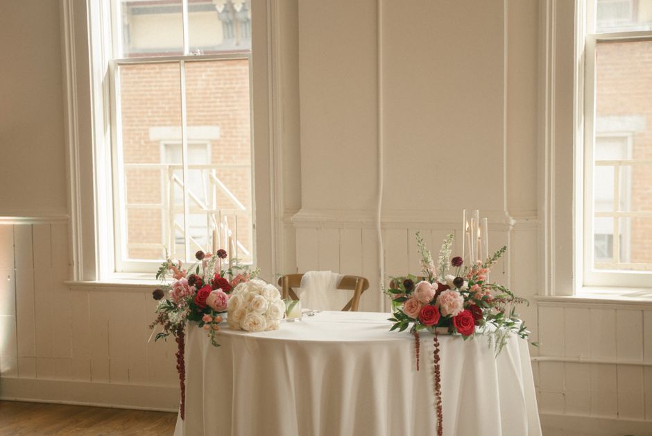 A floral arrangement featuring two matching bouquets placed on a round, white-draped table in a bright, airy room with cream-colored walls and large windows. The bouquets contain pink and red roses, s