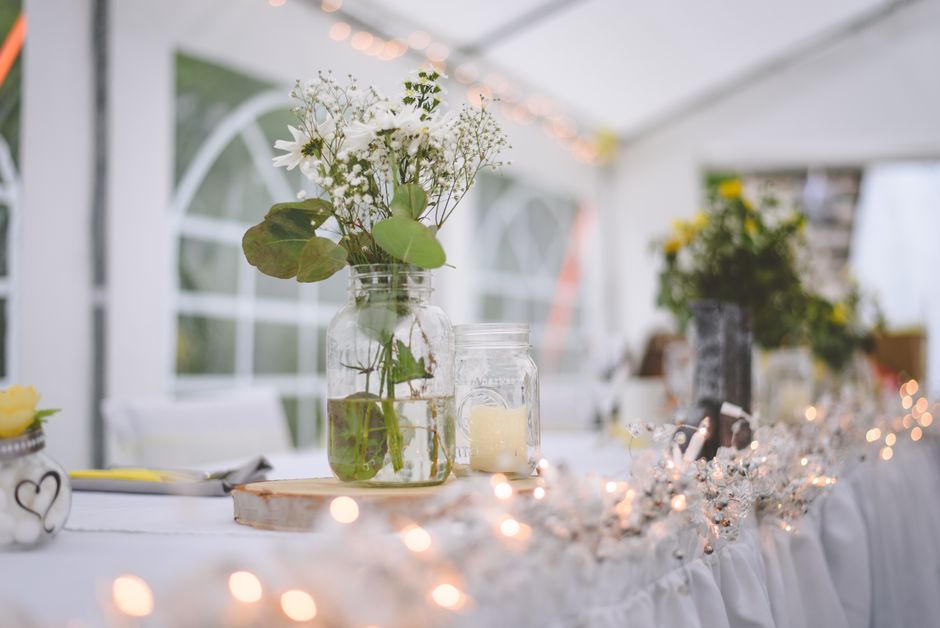 A floral arrangement featuring white daisies, baby's breath, and green leaves placed in a clear glass jar filled with water, positioned on a white tablecloth decorated with small string lights. The ar
