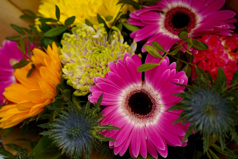 A vibrant floral arrangement featuring large pink gerbera daisies with dark centers, bright yellow-orange sunflowers, pale green and white chrysanthemums, and spiky blue thistle flowers. The bouquet i