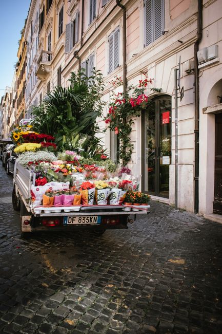 A flower delivery vehicle parked on a cobblestone street in front of a building with cream-colored façade and window shutters, loaded with a vibrant assortment of fresh floral bouquets and potted pla