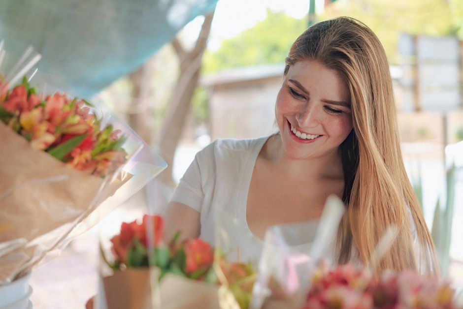 A young woman with long, straight, light brown hair and a cheerful expression is arranging a bouquet of fresh, vibrant flowers. She is wearing a white short-sleeved top and is seated outdoors with nat