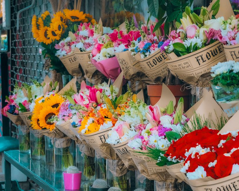 A display of various fresh floral bouquets arranged in wrapping paper with vintage newspaper print, featuring a vibrant assortment of flowers including bright yellow sunflowers, pink lilies, white ros